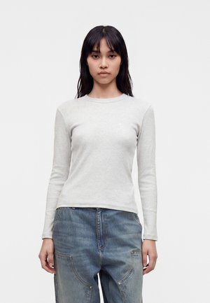 Kvinde med langt mørkt hår iført en lysegrå langærmet t-shirt og løse blå jeans, stående mod en ensartet lys baggrund.