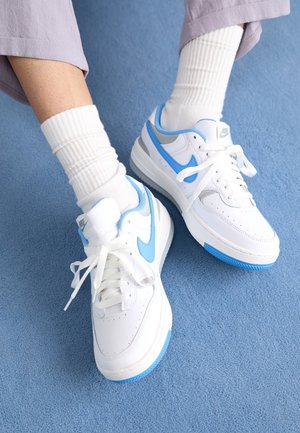 Persoon draagt witte en lichtblauwe Nike-sneakers met witte geribbelde sokken op een blauw tapijt, benen gekruist bij de enkels.