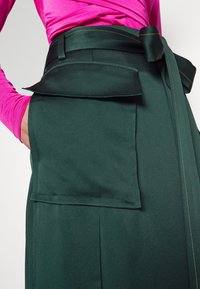 Grön tygkjol med bred midjeband, knytband och två framfickor med kontrasterande sömnad. Slät textur och strukturerad design.
