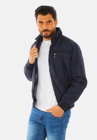 Giacca bomber navy con tasca sul petto con zip, due tasche laterali e polsini a costine. Realizzata in materiale leggero con una texture liscia.