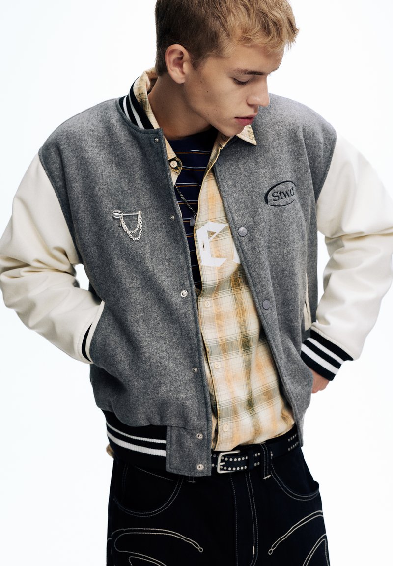 Veste de varsity grise et blanche avec un corps en laine, des poignets rayés et une broche en chaîne argentée. Portée sur une chemise à carreaux aux tons jaunes.