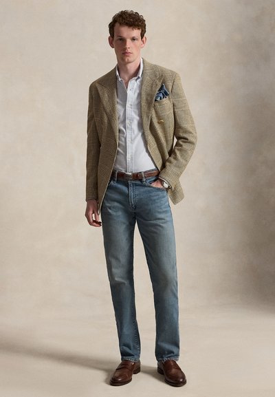Jeune homme portant un blazer à carreaux, une chemise rayée, un jean bleu et des chaussures en cuir marron, debout devant un fond neutre uni.