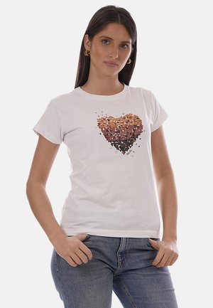 Donna che indossa una maglietta bianca con un cuore formato da piccole farfalle marroni e arancioni, abbinata a jeans blu e orecchini a cerchio dorati.