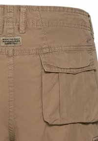 Khaki cargo pants pocket with flap and stitched label reading "Made for daily adventures since 1977" near the waistband.