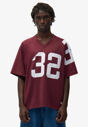 Giovane uomo dalla pelle scura e capelli corti intrecciati, indossa una maglia sportiva color amaranto con il numero 32 bianco e jeans blu larghi, in piedi davanti a uno sfondo semplice.