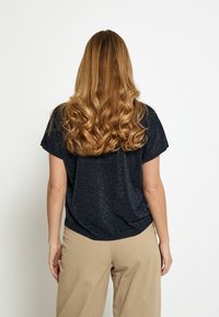 Top a maniche corte di colore blu navy scuro con una texture scintillante; vestibilità comoda. Indossato con pantaloni beige, larghi. Capelli acconciati in morbide onde.