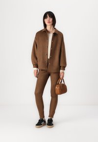 Giacca oversized in camoscio marrone con bottoni, pantaloni slim-fit abbinati, sneaker nere e una piccola borsa a tracolla in pelle marrone testurizzata.