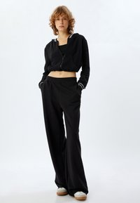 Felpa nera con zip e accenti a righe bianche, abbinata a pantaloni della tuta neri e larghi. Il modello sta in piedi con le mani nelle tasche. Abbigliamento casual.