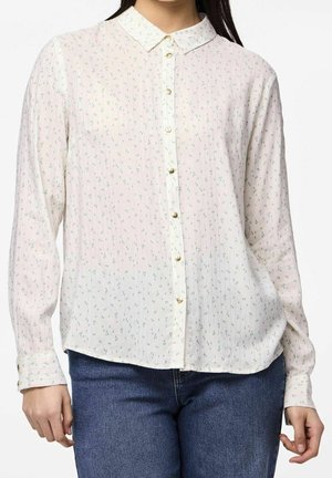 Femme portant un chemisier crème boutonné avec un petit motif floral associé à un jean bleu, montrée du cou aux hanches.