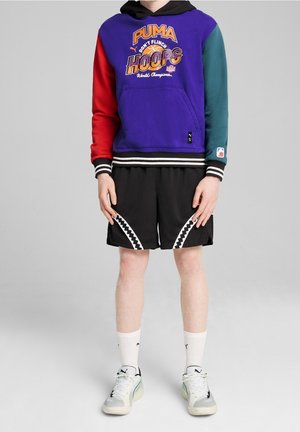 Kolorowa bluza z kapturem PUMA w odcieniach fioletu z czerwonymi i zielonymi rękawami, zdobiona nadrukiem graficznym. Czarne szorty z siateczki z białymi zygzakowymi akcentami.