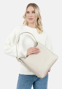 Blonde woman in white sweater holding a large beige leather handbag with adjustable shoulder strap, wearing blue jeans.