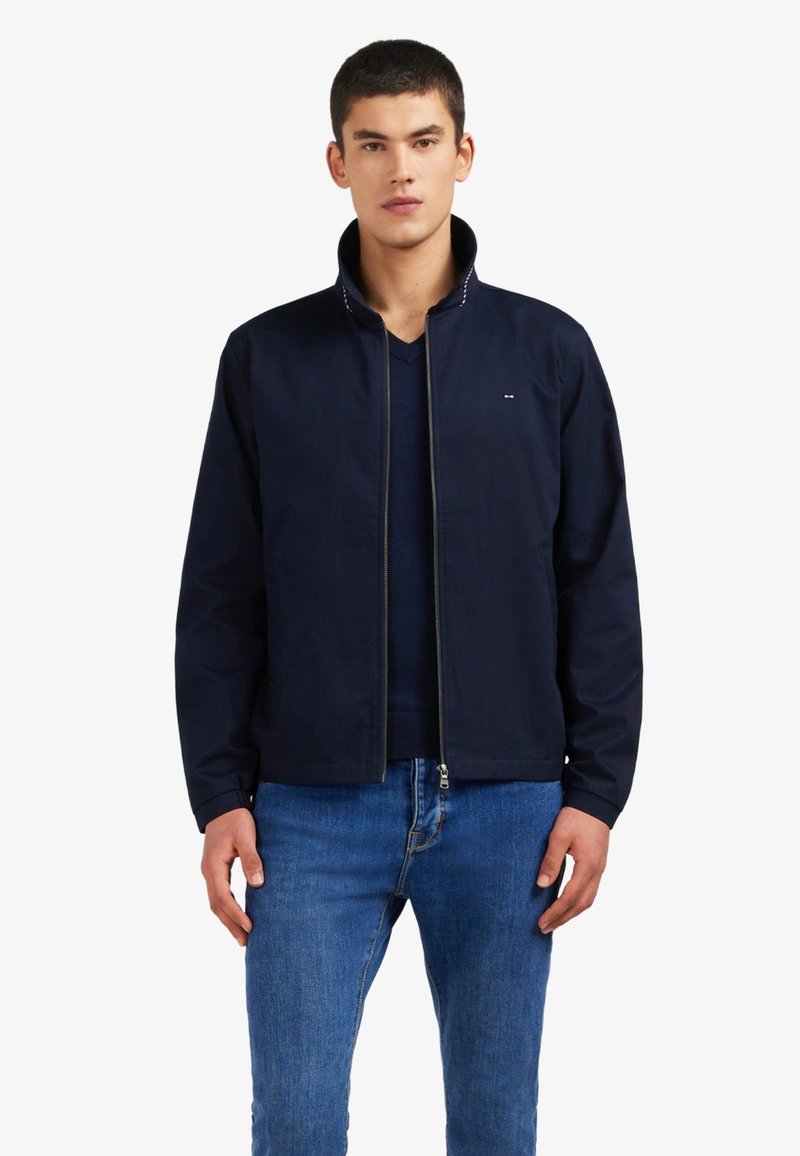 Jeune homme portant une veste zippée bleu marine sur un t-shirt bleu foncé à col en V et un jean bleu, debout face à l’avant sur un fond blanc.