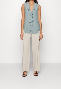 Blouse bleu clair sans manches avec un devant à volants, associée à un pantalon large beige. Des talons noirs à lanières complètent la tenue.