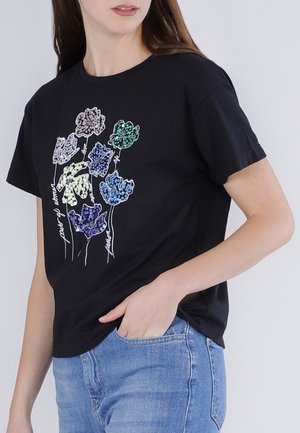 Femme portant un t-shirt noir orné de fleurs en sequins colorés et de texte blanc en écriture cursive, associée à un jean bleu clair, sur un fond uni.