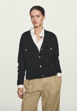 Cardigan nero lavorato a maglia con due tasche frontali, bottoni dorati, polsini a coste, indossato sopra una camicia bianca con colletto. Sfondo semplice.