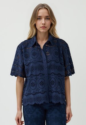 Een vrouw staat met haar gezicht naar de camera, gekleed in een marineblauwe geborduurde blouse met bloemen uitsparingen en korte mouwen.