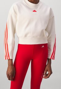 Krémová zkrácená mikina s dlouhými rukávy, která má tři červené pruhy a logo na hrudi. Červené legíny s vysokým pasem a minimálním označením.