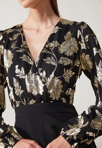Svart blommig blus med gyllene metalliska blommor, djup V-halsringning, texturerad tyg, puffärmar och en figursydd midja.