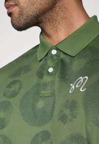 Žalios spalvos polo marškinėliai su tekstūruota apskritimų rašto apdaila, trimis mygtukais ir siuvinėtu balto logotipo ženklu ant krūtinės.