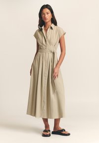Haljina košulja boje kaki s kratkim rukavima, kopčanjem sprijeda i remenom u struku. Lagani materijal dopire do visine lista, s naborima i opuštenim krojem.