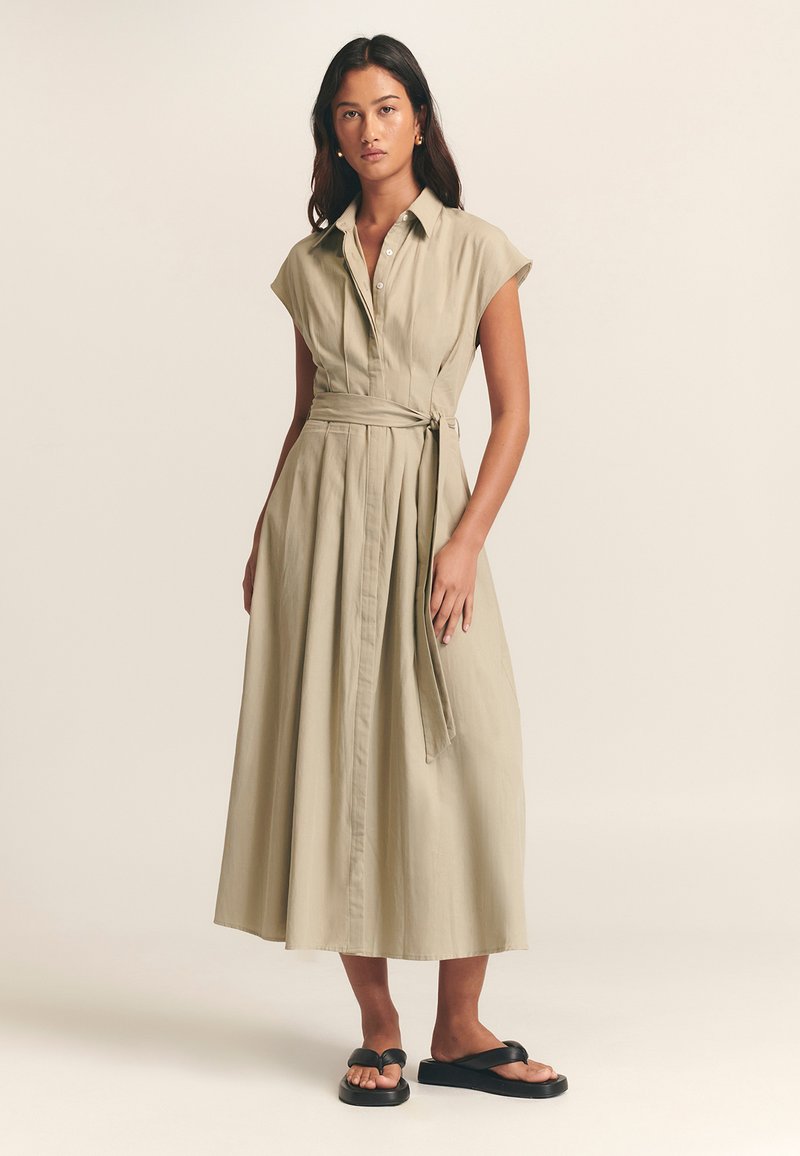 Haljina košulja boje kaki s kratkim rukavima, kopčanjem sprijeda i remenom u struku. Lagani materijal dopire do visine lista, s naborima i opuštenim krojem.