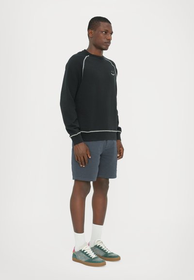 Svart sweatshirt med grå sömmar, kombinerat med marinblå shorts. Modellen bär vita strumpor och gröna sneakers med rosa accenter.