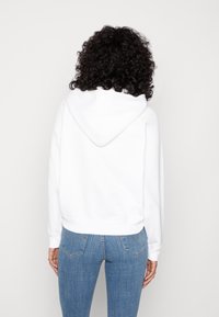 Persona con cabello rizado que lleva una sudadera blanca y jeans azules, de pie frente a un fondo blanco liso.