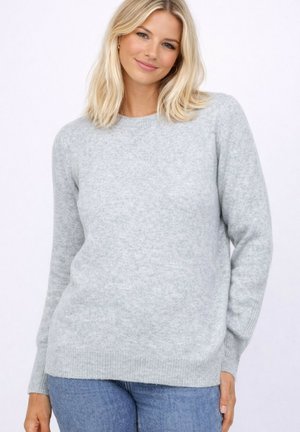 Femme blonde portant un pull en maille gris clair à manches longues et un jean bleu, souriant doucement devant un fond blanc uni.