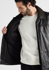 Schwarze Lederjacke mit einem braunen Lammfellkragen, offen getragen, um das schwarze Innenfutter und eine Reißverschlusstasche zu zeigen. Getragen über einem hellen Hemd.