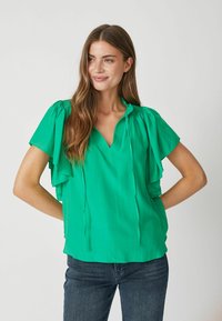 Zelená blúzka s výstrihom do V, s volánkovými detailmi pozdĺž výstrihu a splývavými, vrstvovými krátkymi rukávmi; v kombinácii s tmavými džínsami.