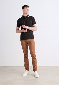 Black polo shirt with white accents, brown slim-fit pants, and white sneakers. Model stands in a neutral background, facing slightly to the side.