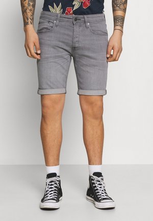 Hombre vestido con pantalones cortos de mezclilla grises arremangados, calcetines blancos, zapatillas negras de caña alta y una camisa con estampado floral, de pie contra un fondo sencillo.