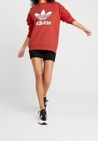 Röd sweatshirt med en vit Adidas-logga och tre ränder, kombinerad med svarta shorts och svarta sportskor. Lågskuren design, avslappnad passform.