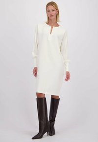 Lange mouw, witte jurk met een keyhole-halslijn en zilveren accenten, gecombineerd met strakke, zwarte stiletto laarzen tot de knie. Soepele stof textuur.