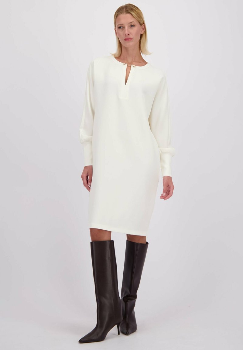 Lange mouw, witte jurk met een keyhole-halslijn en zilveren accenten, gecombineerd met strakke, zwarte stiletto laarzen tot de knie. Soepele stof textuur.