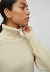 Maglione a collo alto lavorato a maglia color crema con texture a coste e maniche leggermente a sbuffo, che mette in mostra un design aderente.