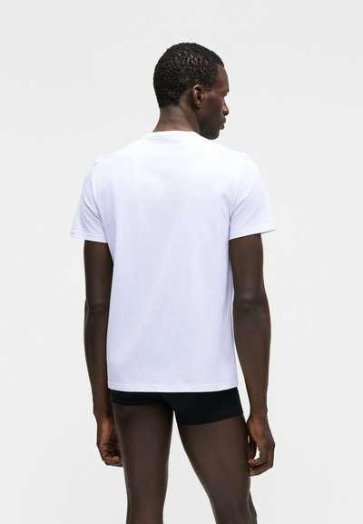 Άνδρας με κοντά σγουρά μαλλιά, με την πλάτη γυρισμένη, φοράει απλό λευκό κοντομάνικο πουκάμισο και μαύρο σορτς, σε φόντο ανοιχτού γκρι χρώματος.