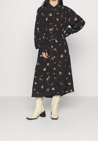 Svart blommig klänning med långa ärmar, krage och midja med bälte. Har orange och vita blommor på ett mjukt tyg. Bärs med beige ankelboots.