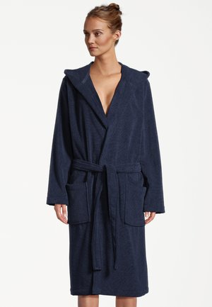 Donna che indossa un accappatoio con cappuccio blu navy con grandi tasche frontali e cintura annodata in vita, in piedi davanti a uno sfondo semplice.