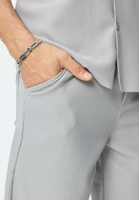Chemise et pantalon rayés verticaux gris, avec un détail de poche. Bracelet en argent au poignet, montrant une texture métallique lisse.