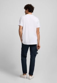 Camiseta blanca de cuello redondo combinada con pantalones cargo navy que cuentan con bolsillos laterales. El modelo lleva zapatillas de lona blancas y sostiene unas gafas de sol en la mano.
