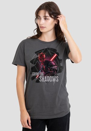 Young woman wearing dark gray t-shirt with red and black character holding a red lightsaber and text "From the Shadows."