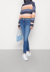 Pestrý pruhovaný rolák v námořnické, oranžové a modré barvě, kombinovaný s úzkými modrými džíny a bílými teniskami. Drží světle modrou kabelku.