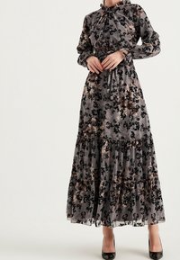 Pitkähihaista maksimekkoa, joka on valmistettu läpinäkyvästä harmaasta kankaasta ja jossa on mustia ja hillittyjä sävyjä sisältävä kukkakuosi. Mekossa on röyhelöillä varustettu kerroksellinen helma ja korkea kaulus.