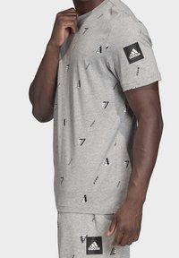 Szara koszulka z bawełny z nadrukiem małych czarnych logo Adidas w całym wzorze. Teksturowany materiał z okrągłym dekoltem i krótkimi rękawami.