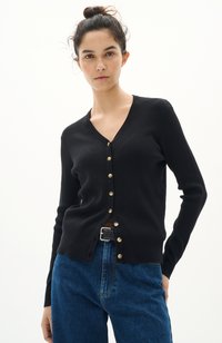 Svart ribbad cardigan med V-hals, guldknappar fram och slim fit. Kombination med högmidjade blå denimjeans.