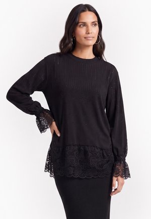 Frau trägt ein schwarzes Langarmshirt mit Spitzenbesatz an Bündchen und Saum, kombiniert mit einem schwarzen Ripprock, steht mit einer Hand in der Hüfte.