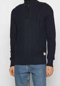 Marinblå kabelstickad tröja med hög krage och halv dragkedja. Har texturerade mönster samt ribbade ärmslut och nederkant. Bärs över ljusblå jeans.