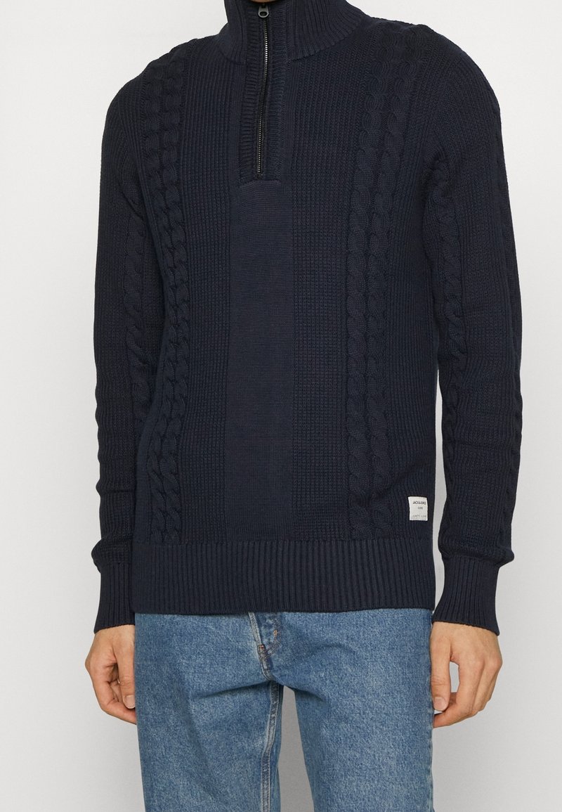 Marinblå kabelstickad tröja med hög krage och halv dragkedja. Har texturerade mönster samt ribbade ärmslut och nederkant. Bärs över ljusblå jeans.