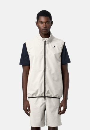 Modello maschile che indossa un gilet senza maniche color beige chiaro con cerniera sopra una maglietta nera a maniche corte e pantaloncini beige coordinati, in piedi davanti a uno sfondo neutro.